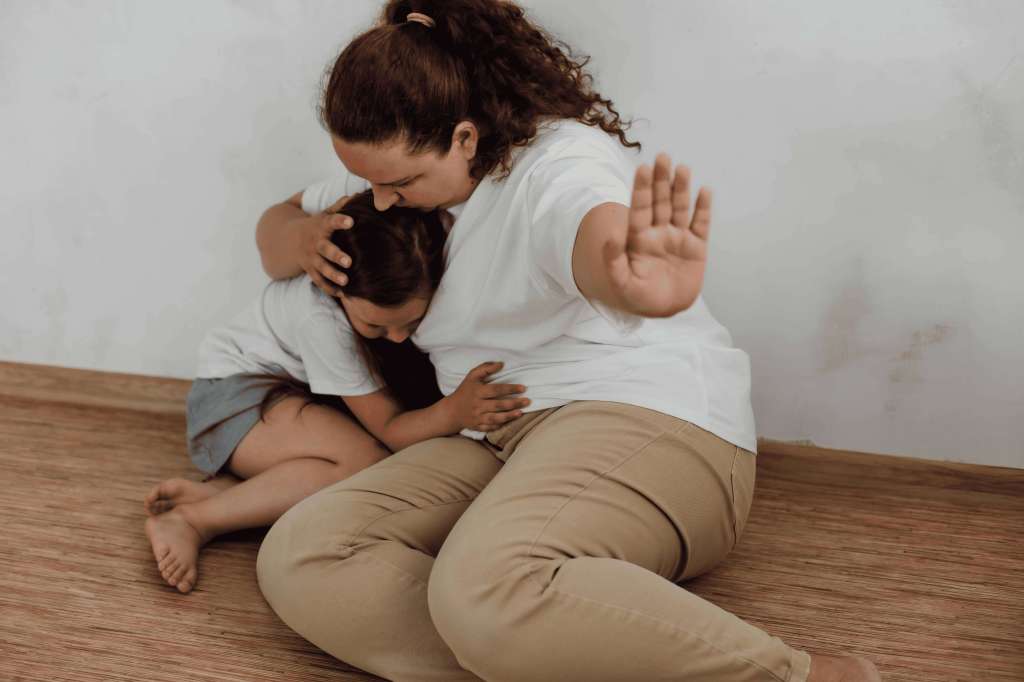 Una madre abraza fuertemente a su hija mientras extiende una mano en señal de detención, simbolizando protección y la vulnerabilidad infantil.