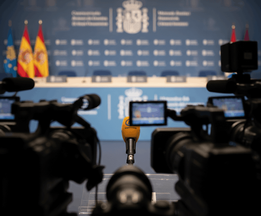 Un micrófono de prensa solitario en primer plano, rodeado por las siluetas de cámaras de televisión desenfocadas. Al fondo, un panel institucional con banderas y logotipos gubernamentales en una sala de prensa vacía, transmitiendo una sensación de puesta en escena política.