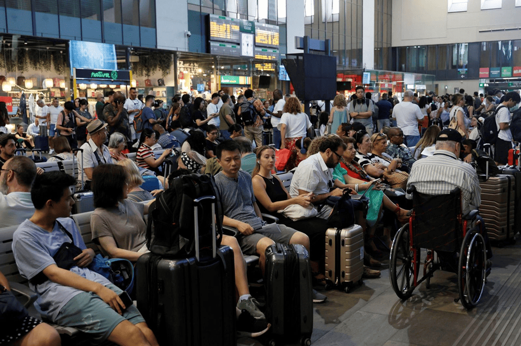 Estación de tren con cientos de personas sentadas en el suelo del andén, maletas amontonadas en un ambiente de abandono.