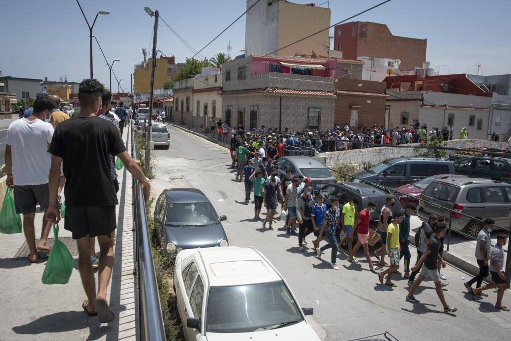 Un grupo numeroso de personas camina en fila por una calle asfaltada junto a coches aparcados y casas en Ceuta.