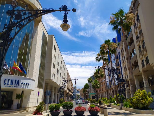 Vista urbana de una avenida céntrica en Ceuta con el Hotel Puerta de África, palmeras, jardineras y farolas ornamentales bajo un cielo despejado.