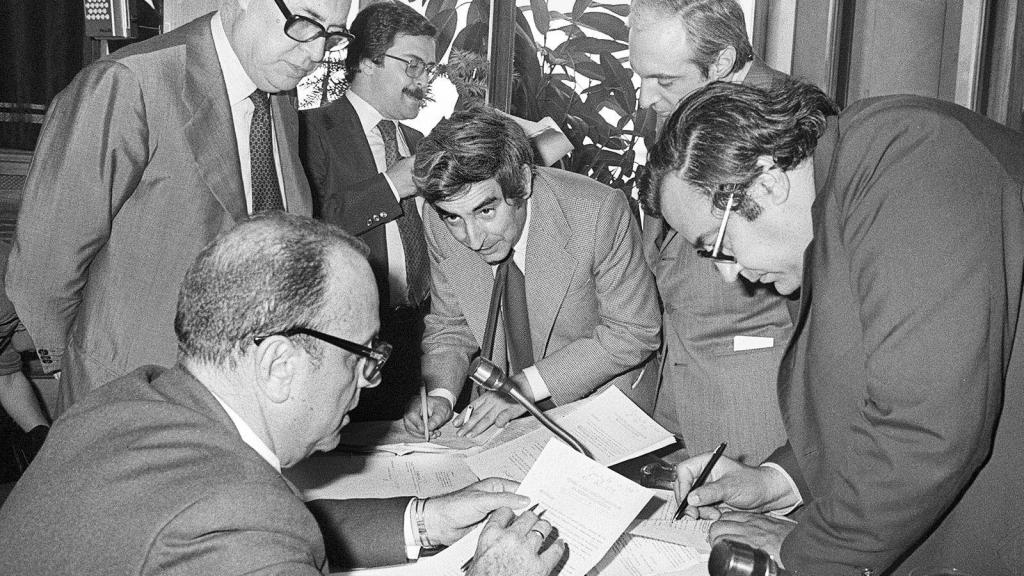 Fotografía histórica en blanco y negro de políticos españoles firmando los Pactos de la Moncloa en 1977, reunidos alrededor de una mesa examinando documentos.