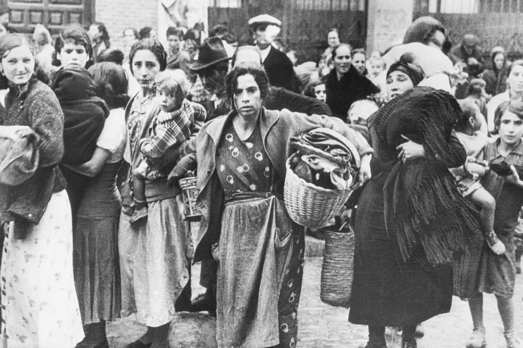 Fotografía histórica en blanco y negro de mujeres, niños y familias evacuadas caminando por la calle cargando cestas y mantas durante la Guerra Civil Española.