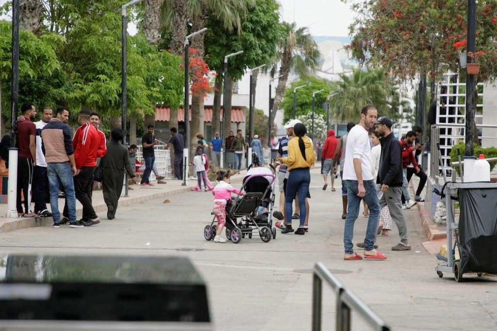 Personas paseando y conversando en un paseo peatonal arbolado en Ceuta, con niños y carritos de bebé.